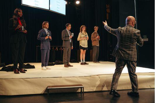Man pointing to 5 candidates on stage, some dressed in business and some dressed casually
